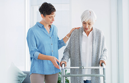 Female caregiver helping an older woman walk with a mobility walker indoors
