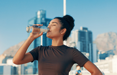 Woman drinking water outdoors with city buildings in the background on a sunny day